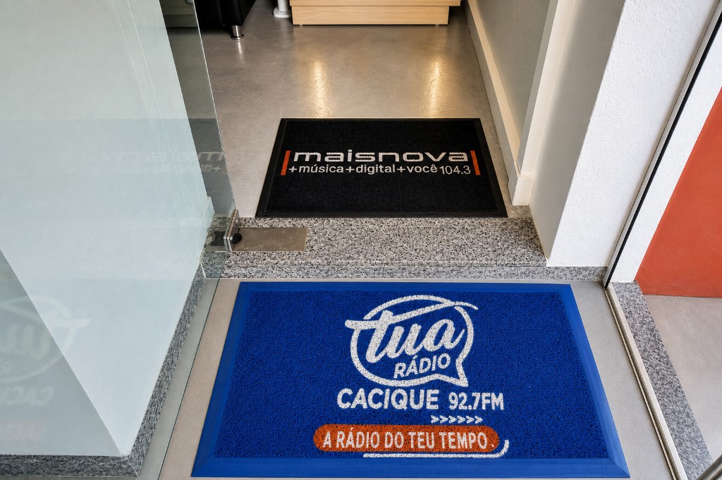 Capachos personalizados das rádios Maisnova 104.3 e Cacique 92.7FM instalados na entrada sobre piso premium, com identidade das emissoras em destaque.