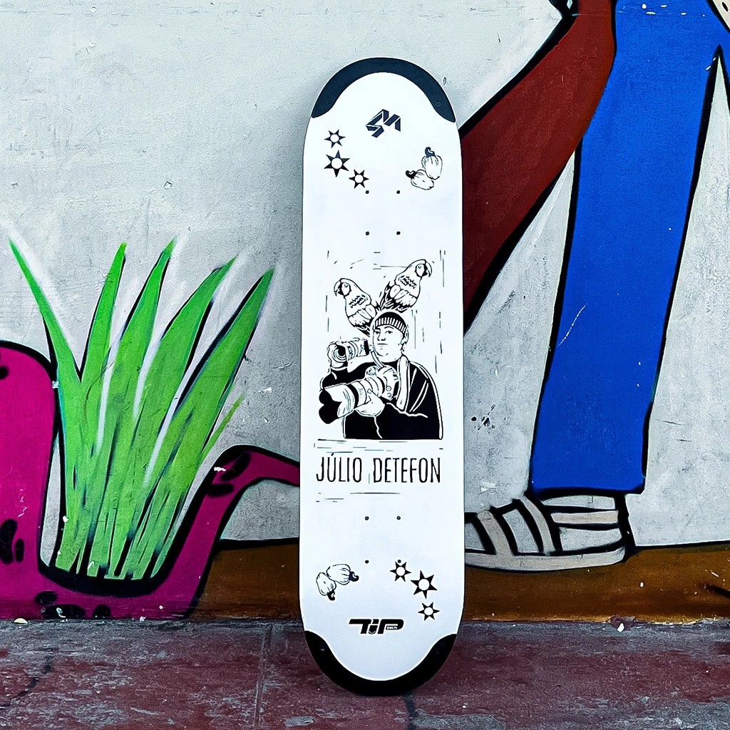 Shape de skate branco da marca 4Msb, pro model Júlio Detefon, encostado em uma parede grafitada e destacando a tecnologia TIPtech preta no nose e no tail.
