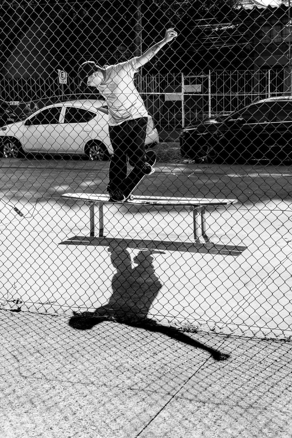 Fotografia em preto e branco de um skatista de rua executando uma manobra de borda deslizando a parte frontal do skate (noseslide) em um banco de praça, visto através de uma grade de arame. Manobras de impacto em obstáculos urbanos exigem extrema resistência estrutural da madeira. O uso de um shape durável com a tecnologia TIPtech da 4Msb by Skateovation garante que a prancha suporte essa pressão extrema. Essa engenharia de skate absorve os choques e protege contra o desgaste rápido, garantindo um pop duradouro sem alterar o peso ou o estalo seco do skate. Essa performance elevada evita a quebra precoce e promove um consumo consciente no skate, alinhado à sustentabilidade do esporte.