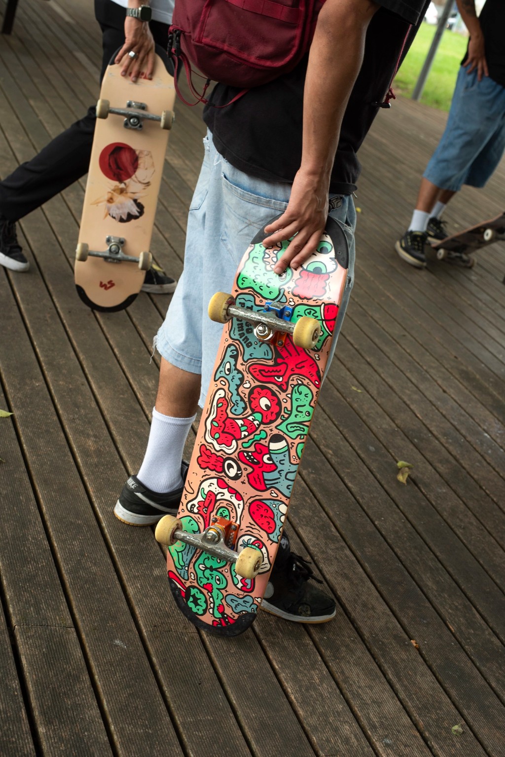 Skatista em pé segurando pela lateral um shape da 4Msb (modelo Tiago Picomano Doodle) com gráficos coloridos. Nas extremidades da prancha (nose e tail), destacam-se os enxertos escuros da tecnologia TIPtech. Essa engenharia de skate absorve os impactos diários, mantendo a tensão estrutural do concave e garantindo um pop duradouro. Ao proteger a madeira contra o desgaste do razor tail sem alterar o peso ou o estalo seco natural do shape, o equipamento entrega alta performance e promove o consumo consciente no skate, diminuindo o descarte prematuro.