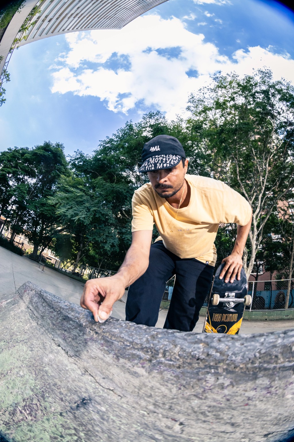 Fotografia com lente grande angular (fisheye) de um skatista agachado passando parafina na borda de uma transição de concreto em uma pista ao ar livre, preparando o pico para a sessão. Em sua mão esquerda, ele segura o seu skate, deixando o gráfico do modelo Tiago Picomano visível. Passar parafina ajuda a deslizar nas manobras, mas o atrito constante em obstáculos de concreto é o maior causador do desgaste acelerado e da delaminação de pranchas tradicionais. Para suportar essa fricção pesada do street, o skatista utiliza um shape com a tecnologia TIPtech da 4Msb by Skateovation. Essa engenharia de skate atua protegendo o nose e o tail, entregando um pop duradouro e estendendo a vida útil do equipamento em até 3x. O reforço estrutural não altera o peso do skate e garante que o estalo continue nítido e seco, consolidando-se como o melhor shape para manobras e um pilar para o consumo consciente no skate.