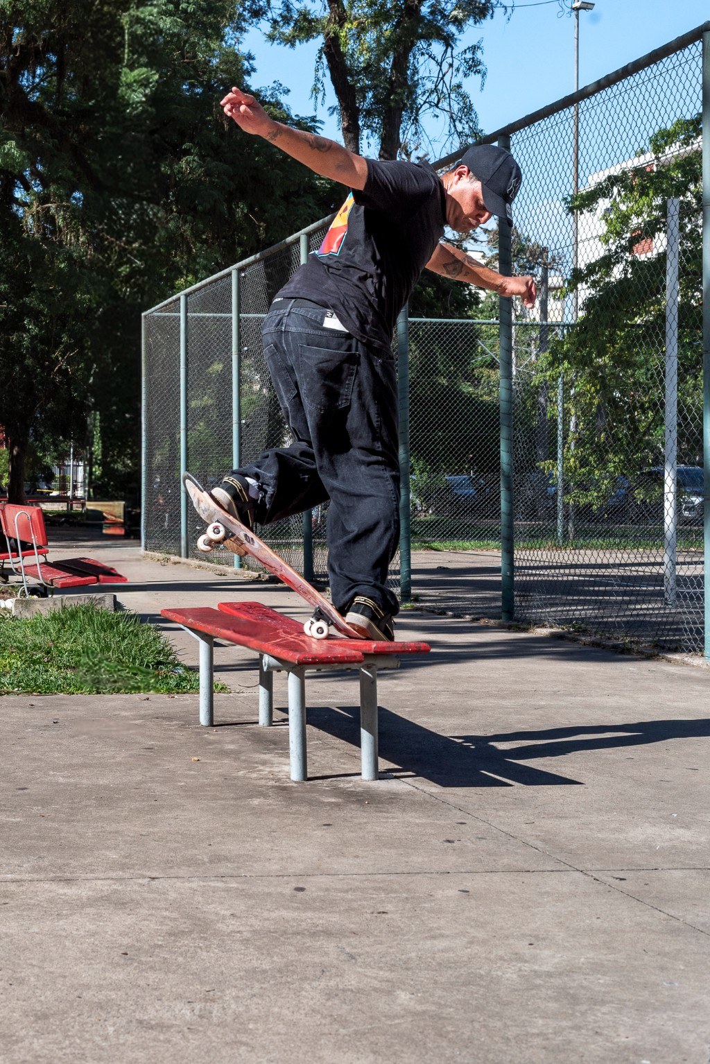 Skatista vestindo preto (bon&eacute;, camiseta, jeans) realizando manobra de 5-0 stall no truck traseiro em um banco de parque vermelho de metal. Cerca de arame e &aacute;rvores ao fundo sob luz solar brilhante.