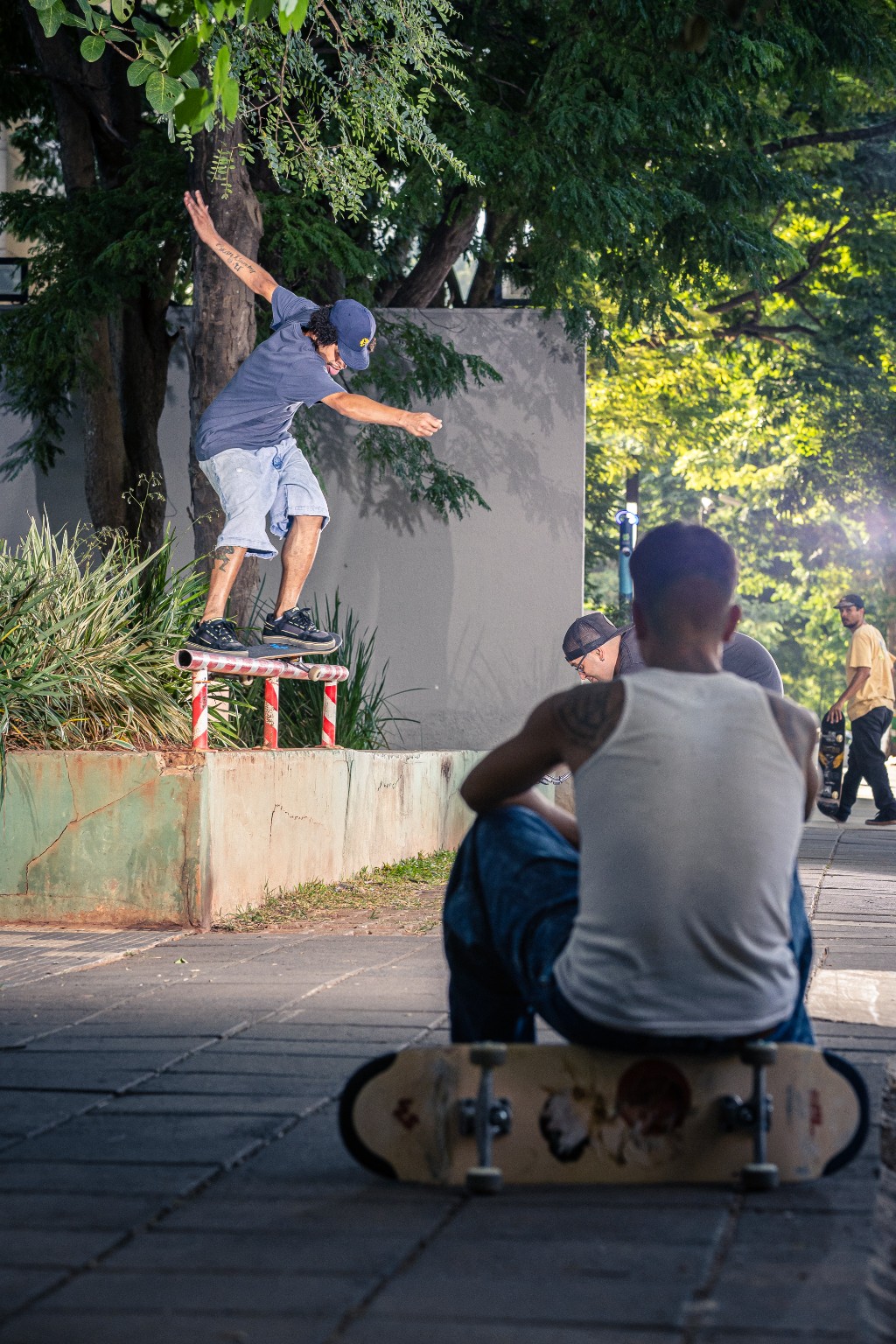 Foto vertical de um skatista (camisa azul, bermuda jeans, bon&eacute; azul) realizando uma manobra crooked grind em um corrim&atilde;o listrado em uma rua urbana. Outro skatista observa sentado em seu skate no primeiro plano. &Aacute;rvores ao fundo.