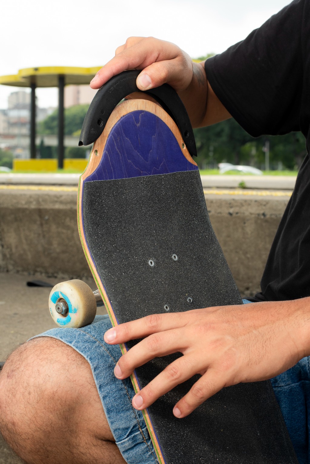 Close-up detalhado mostrando as m&atilde;os de um skatista instalando a ponteira preta TIPtech na borda roxa de um shape de skate 4Msb. Griptape e rodinhas vis&iacute;veis. O skate repousa sobre o joelho do skatista.