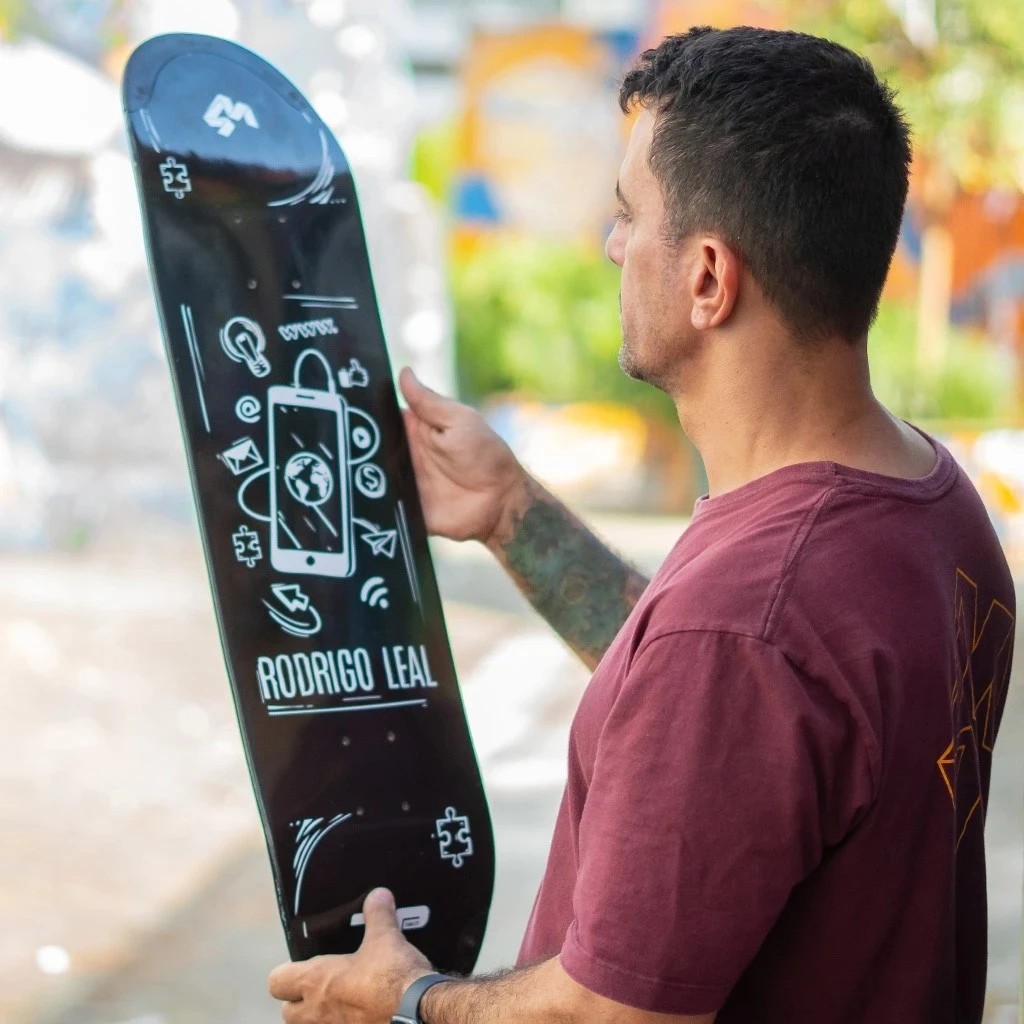 Skatista segurando verticalmente um shape 4Msb preto, pro model Rodrigo Leal, com gráficos em branco. Ele observa os detalhes e o acabamento da prancha nova, pronta para entregar alta performance e durabilidade na sessão.