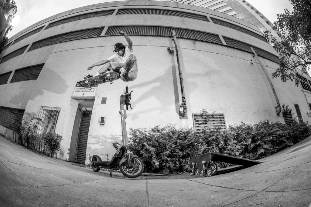 Fotografia urbana em preto e branco com lente olho de peixe. Um skatista marreta um ollie alto por cima de um patinete elétrico estacionado na calçada, mostrando a resistência e o pop do seu shape 4Msb nas ruas.