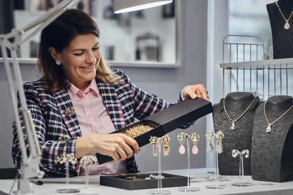 Mulher organizando semijoias em expositores sobre uma mesa de trabalho. Brincos e colares s&atilde;o posicionados com cuidado em suportes, enquanto outros permanecem em caixas abertas. A cena representa os bastidores do lan&ccedil;amento, com foco na apresenta&ccedil;&atilde;o visual dos produtos.