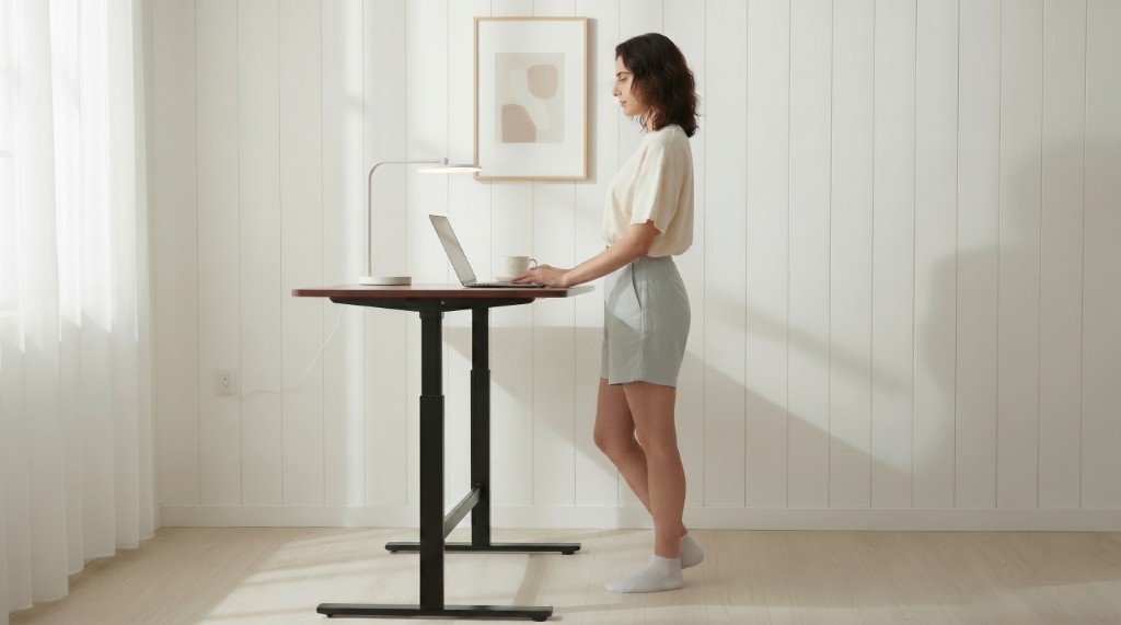 Pessoa trabalhando em pé utilizando uma mesa ergonômica com regulagem de altura e notebook em ambiente minimalista.