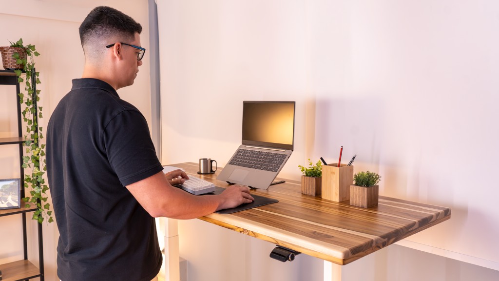 Homem de aproximadamente 1,90m utilizando a mesa F50 em sua altura máxima, trabalhando em pé com notebook em um ambiente minimalista e moderno.