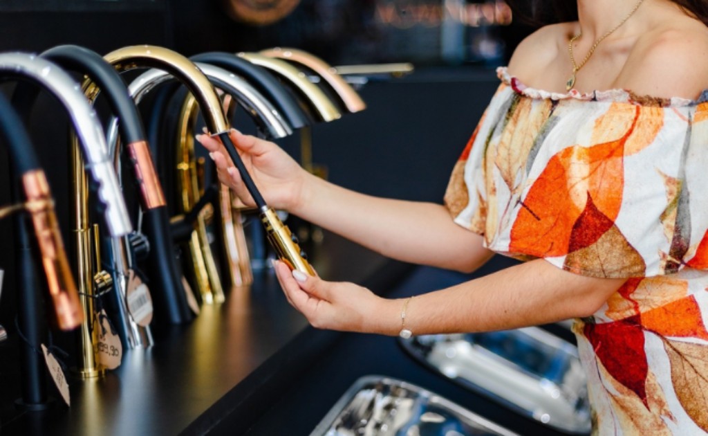 Imagem interna do expositor de torneiras da loja Bruno Acabamentos, uma cliente est&aacute; testando o cano extens&iacute;vel de uma torneira de cozinha dourada! Venha ter essa experi&ecirc;ncia em nossa loja tamb&eacute;m!