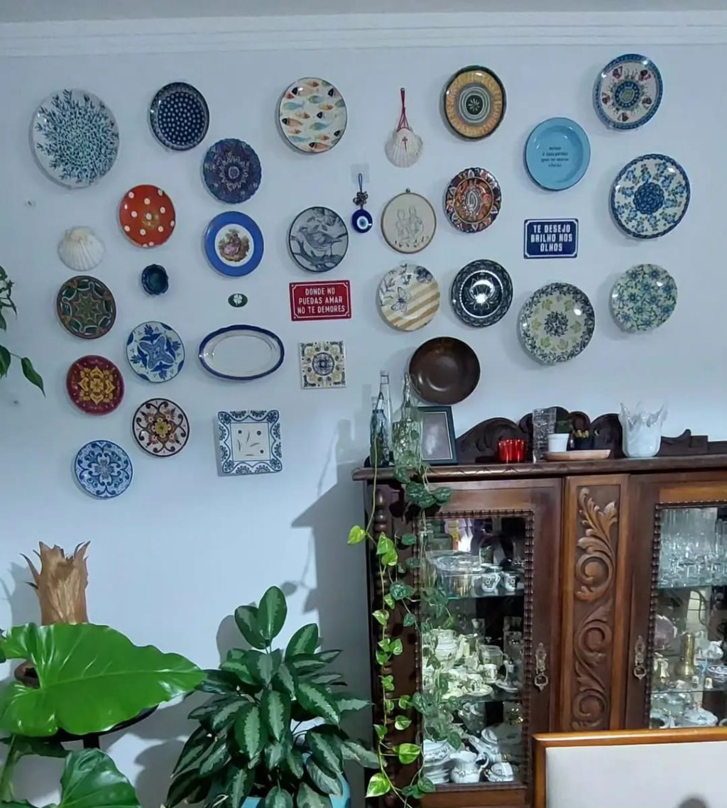 Decora&ccedil;&atilde;o afetiva em sala de jantar com pratos antigos de diversos estilos, plantas ornamentais e cristaleira vintage de madeira ao fundo.