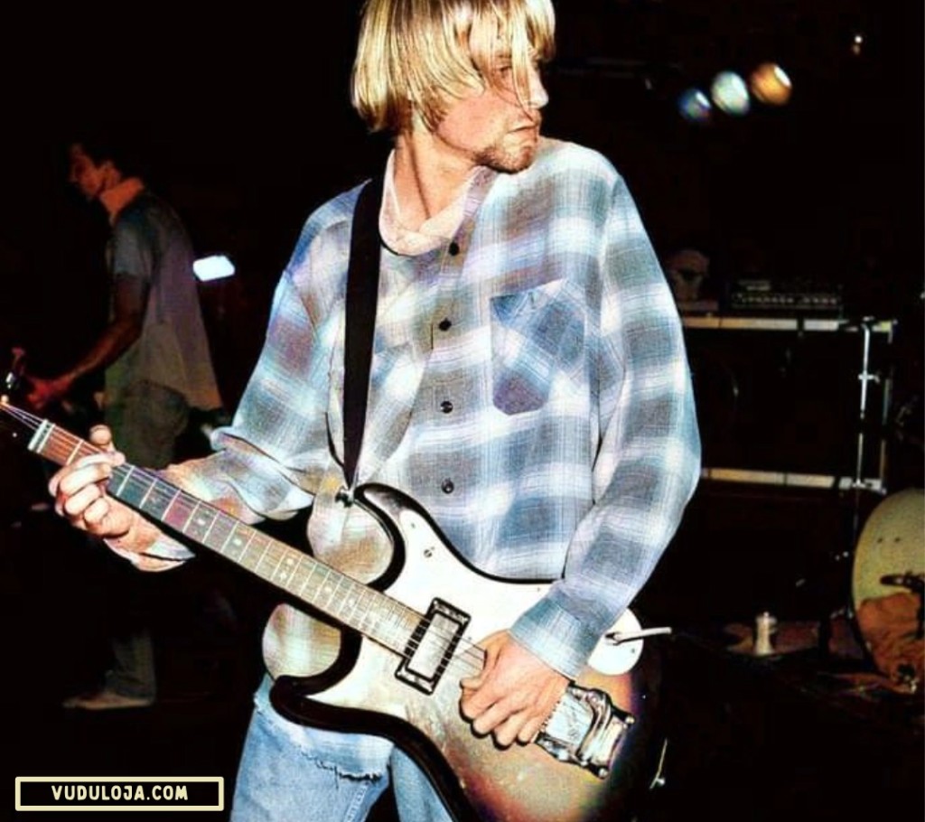 Foto colorida de Kurt Cobain tocando guitarra e vestindo uma camisa work flanela