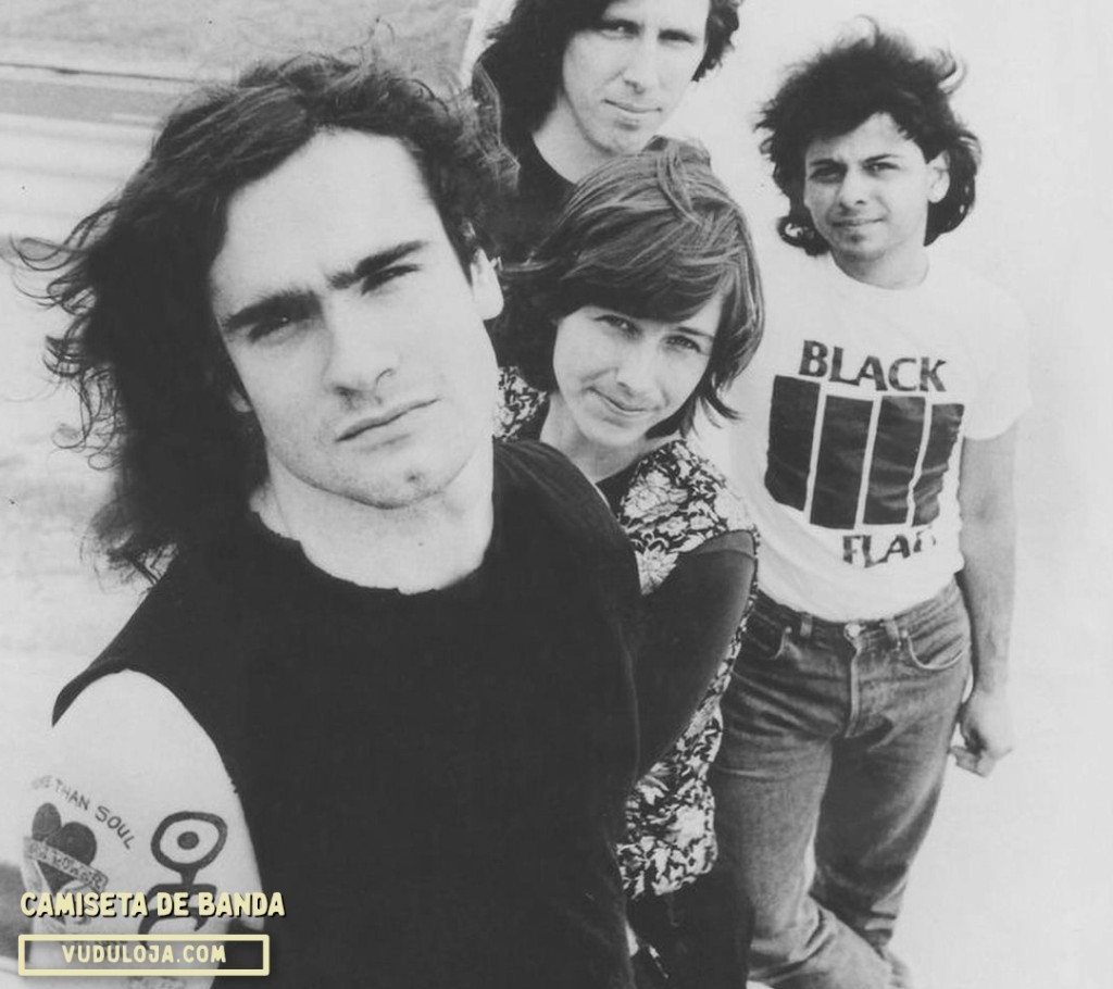 Foto em preto e branco com os integrantes da banda de punk rock Black Flag, destacando um rapaz ao fundo usando uma camiseta de banda branca com o ic&ocirc;nico logotipo de quatro barras pretas da banda.