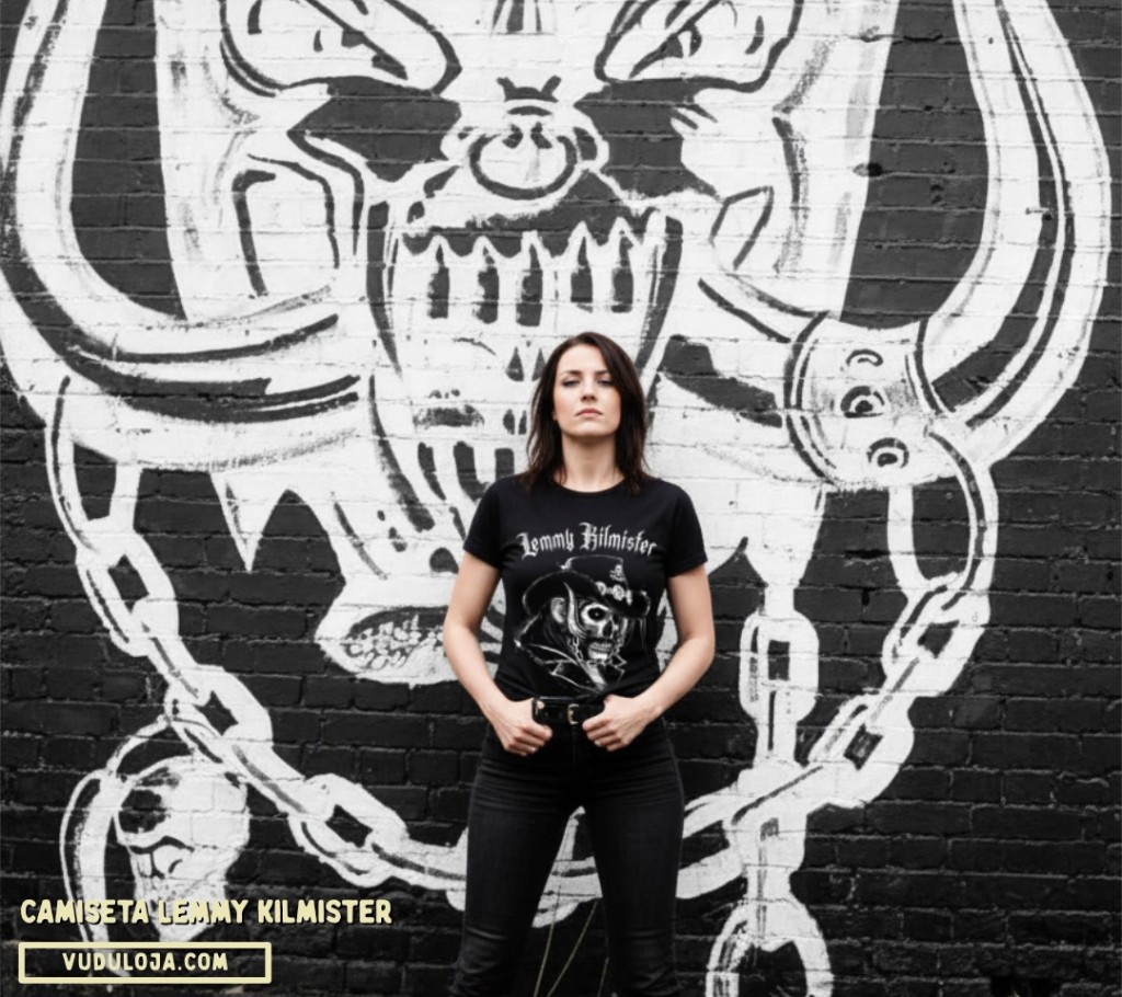 Mulher de cabelos escuros posando &agrave; frente de um muro com grafite do Warpig, usando cal&ccedil;a preta e uma camiseta de banda preta com estampa de Lemmy Kilmister.