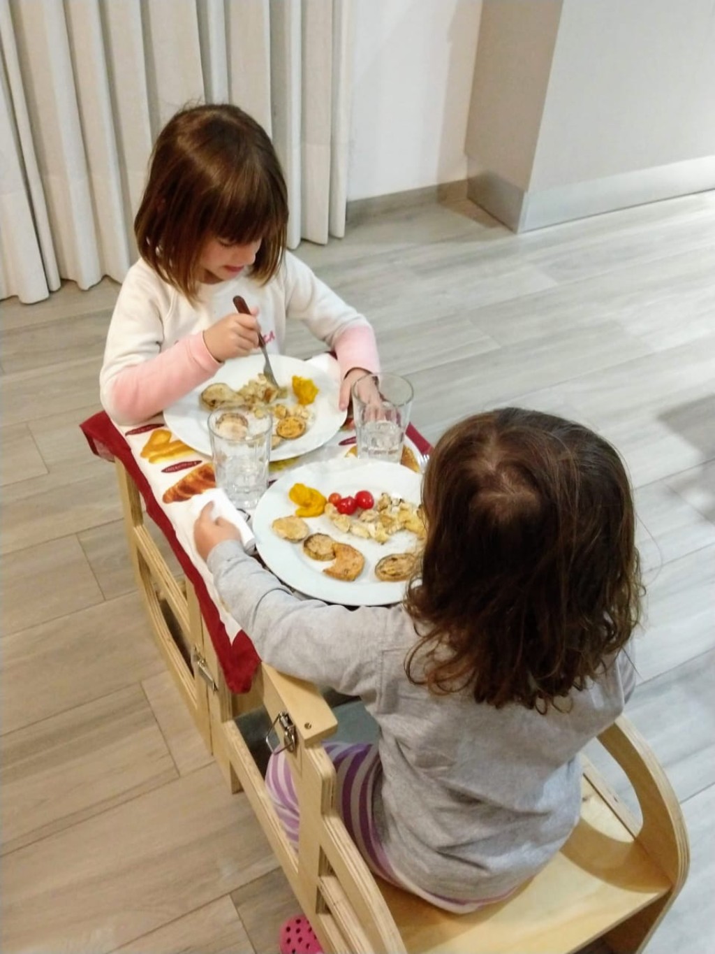 Dos hermanas compartiendo la mesa familiar, sentadas sobre la Torre de Aprendizaje Montedorf transformada en mesa y banco