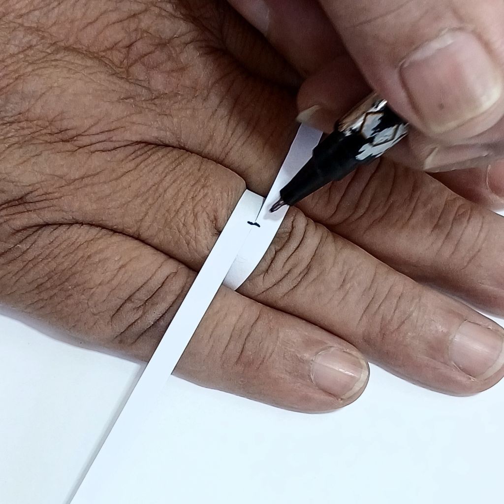 Tutorial passo a passo de como medir o tamanho do dedo em casa para an&eacute;is e alian&ccedil;as. A imagem mostra uma pessoa usando uma tira de papel e caneta para marcar a circunfer&ecirc;ncia do dedo.