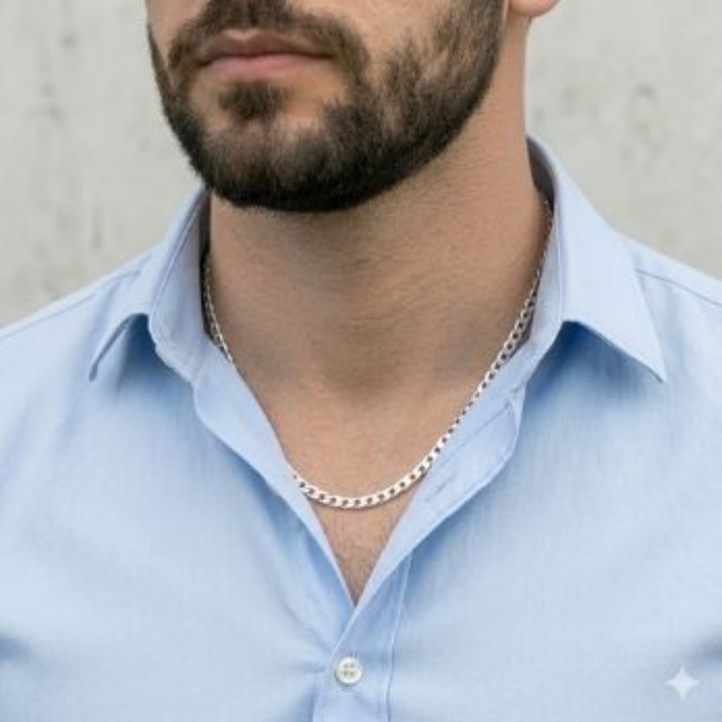 Close-up t&eacute;cnico padronizado do torso de um homem com barba, vestindo uma camisa social azul clara e maci&ccedil;a. O fundo &eacute; um muro de concreto urbano claro. Ele usa uma corrente Piastrine Fita em Prata 925 maci&ccedil;a e achatada, com 60cm de comprimento. A corrente &eacute; usada por baixo do colarinho e aparece sutilmente atrav&eacute;s dos dois bot&otilde;es abertos superiores, com caimento suave e brilhante.
