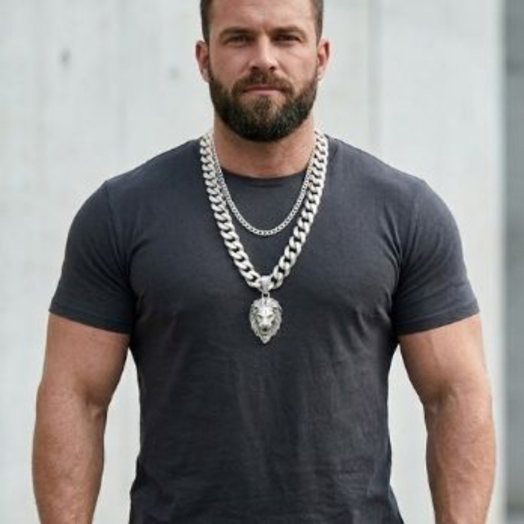 Estudo comparativo padronizado em plano aberto do man com porte robusto, vestindo a camiseta de gola careca na cor carv&atilde;o escura. O fundo &eacute; um muro de concreto urbano claro. Ele demonstra a t&eacute;cnica de 'layering' correto com duas correntes em Prata 925: uma Grumet maci&ccedil;a e robusta (8mm) de 70cm, que serve como base com um pingente de le&atilde;o polido, e uma corrente Piastrine mais fina (2mm) de 50cm, posicionada em um 'degrau' acima, criando uma composi&ccedil;&atilde;o de estilo ousada e proporcional.