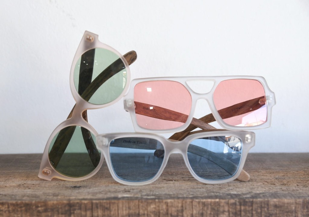 anteojos de madera y acetato marca Nomade con lentes tintados en colores verde, tojo y celeste sobre repisa de madera con fondo blanco