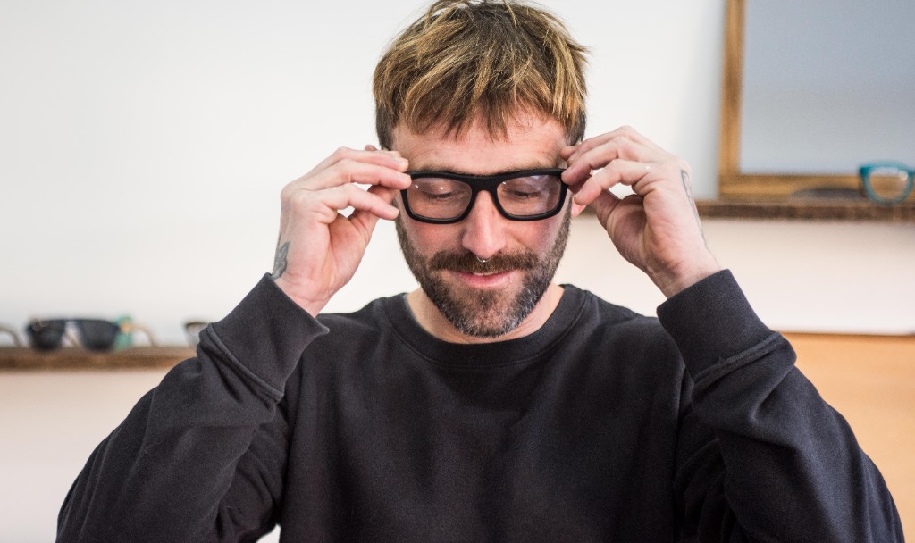 hombre con pelo corto y rubio poniendose anteojos con armazon negro y patillas de madera, luce un sweater color negro en interior con luz natural