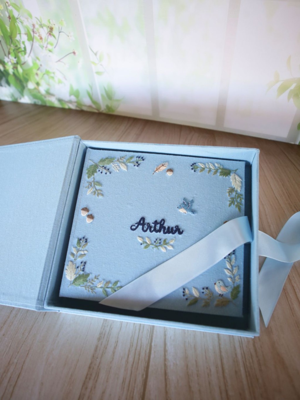 Livro do bebê personalizado com nome bordado à mão em capa de linho, dentro da caixa artesanal, mostrando detalhes do acabamento