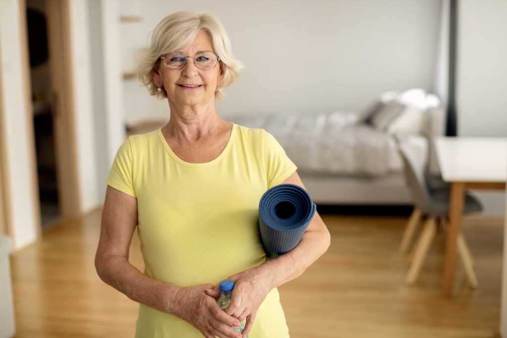 Mujer mayor feliz sosteniendo una colchoneta de ejercicios y agua en casa, demostrando que mantener una vida activa y saludable es posible con EPOC.