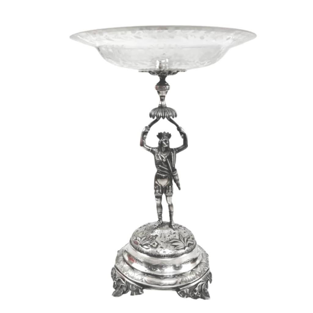 Centro de mesa em prata de lei contrastada, ricamente cinzelada e fundida, produzido em Portugal (com marca localizando no Porto) no s&eacute;culo XIX, acompanhado por prato em cristal lapidado &agrave; m&atilde;o.  A base escult&oacute;rica, trabalhada em alto n&iacute;vel t&eacute;cnico, apresenta figura em postura cl&aacute;ssica sustentando o corpo superior da pe&ccedil;a, revelando o virtuosismo da ourivesaria portuguesa oitocentista. O trabalho minucioso da prata, com volumes bem definidos e detalhes ornamentais, dialoga com a transpar&ecirc;ncia e a precis&atilde;o do cristal lapidado, criando um conjunto de forte presen&ccedil;a visual e equil&iacute;brio formal.  Uma pe&ccedil;a de car&aacute;ter decorativo e cerimonial, representativa da tradi&ccedil;&atilde;o europeia de objetos de mesa e centro, pensada para ambientes que valorizam hist&oacute;ria, t&eacute;cnica e perman&ecirc;ncia.
