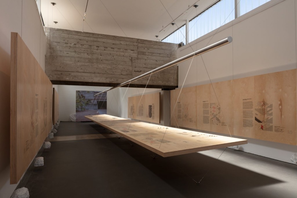 . Exposição "Utopias de Proximidade", Pavilhão do Brasil, Bienal de Veneza. A montagem destaca a expografia suspensa com painéis de madeira e cabos de aço, dialogando com a estrutura de concreto bruto do pavilhão.