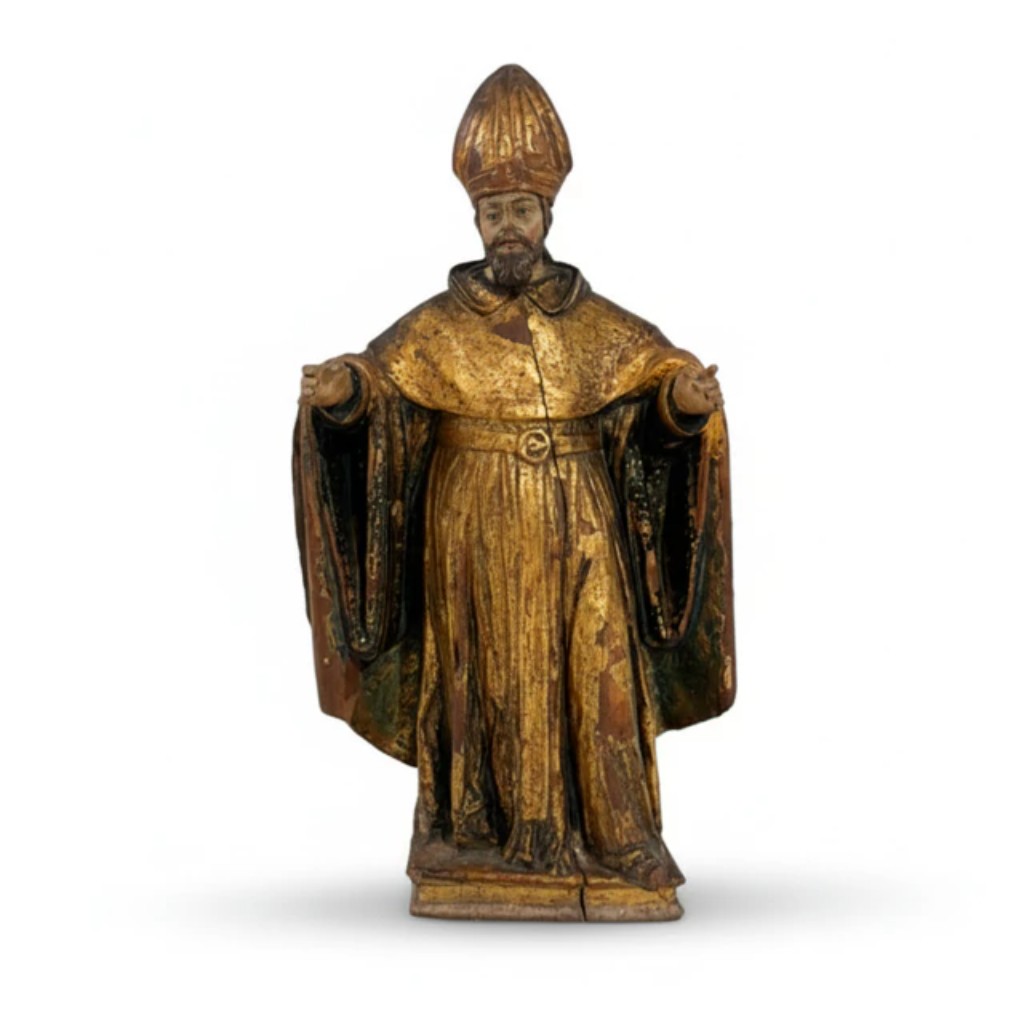 Escultura em madeira entalhada, policromada e dourada representando um santo bispo, figurado com mitra e amplas vestes lit&uacute;rgicas ricamente douradas. A imagem apresenta bra&ccedil;os abertos em gesto de acolhimento e b&ecirc;n&ccedil;&atilde;o, caracter&iacute;stica recorrente da imagin&aacute;ria devocional barroca. O tratamento das vestes, com amplos panejamentos e aplica&ccedil;&atilde;o de douramento, evidencia a tradi&ccedil;&atilde;o da escultura religiosa portuguesa, profundamente ligada &agrave; produ&ccedil;&atilde;o destinada a igrejas, capelas e orat&oacute;rios particulares. Pela iconografia e atributos, a figura &eacute; provavelmente identificada como S&atilde;o Paulo. Portugal, s&eacute;culo XVIII.  