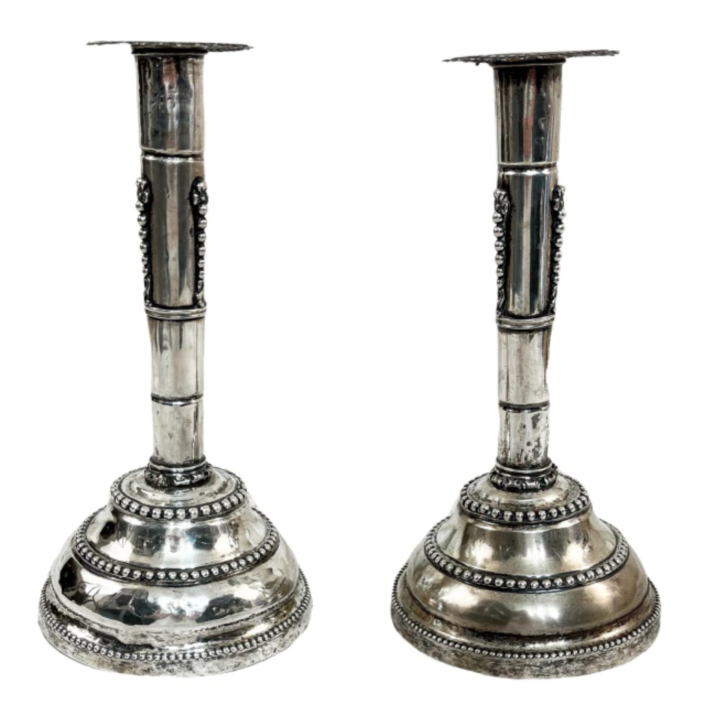 Par de tocheiros em prata de lei, elaborados com as t&eacute;cnicas de batimento, repuxo, cinzelamento e perolado, que conferem riqueza de detalhes e refinamento &agrave; pe&ccedil;a. O batimento e o repuxo moldam a prata, criando volumes harmoniosos, enquanto o cinzelamento adiciona ornamenta&ccedil;&otilde;es esculpidas com precis&atilde;o e o perolado destaca delicados relevos decorativos. Apresentam almas em madeira na parte inferior, refor&ccedil;ando sua estrutura. Produzidos no Brasil, no estilo e &eacute;poca de Dona Maria, no final do s&eacute;culo XVIII, refletem a sofistica&ccedil;&atilde;o e a tradi&ccedil;&atilde;o da ourivesaria luso-brasileira da &eacute;poca.