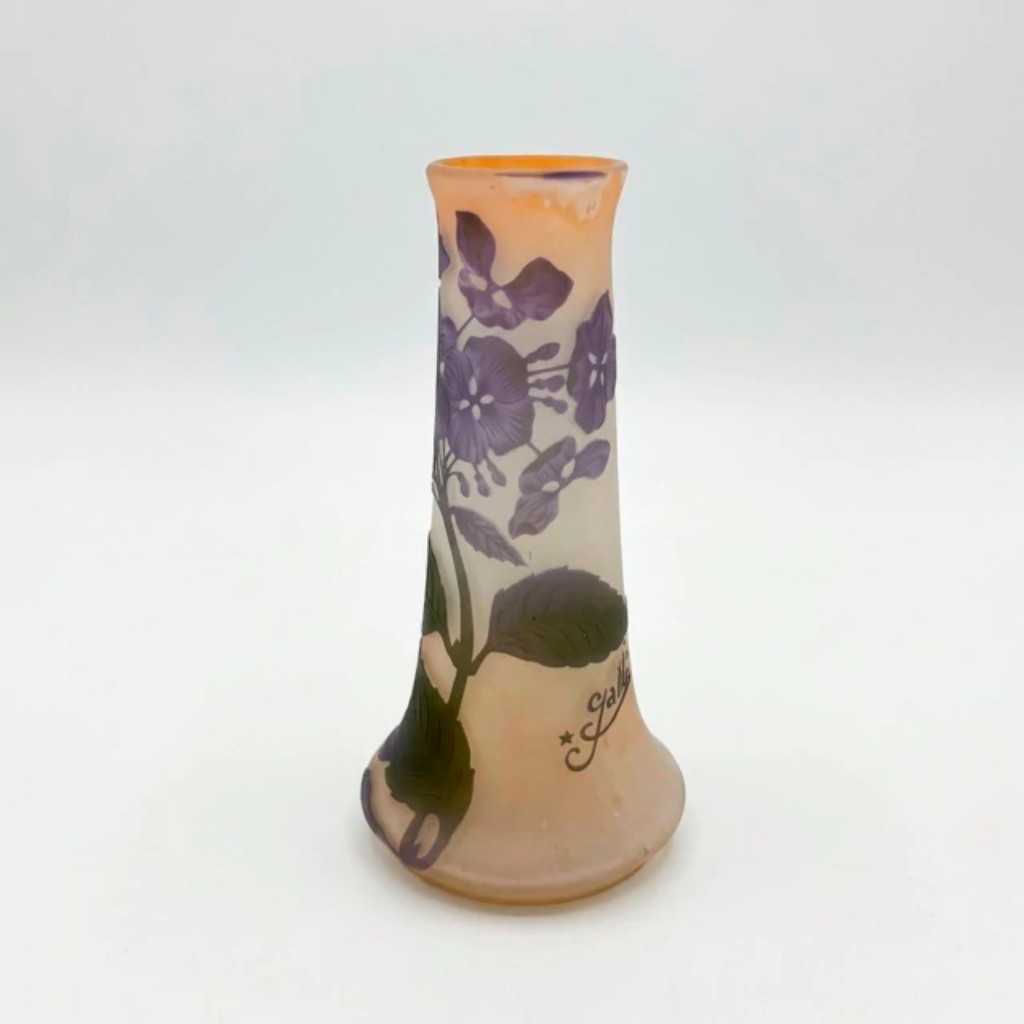 Vaso em vidro cameo produzido pela Gall&eacute; &mdash; manufatura fundada por &Eacute;mile Gall&eacute; em Nancy, Fran&ccedil;a, no final do s&eacute;culo XIX e uma das principais express&otilde;es do movimento Art Nouveau. A t&eacute;cnica cameo consiste na sobreposi&ccedil;&atilde;o de camadas de vidro colorido que s&atilde;o ent&atilde;o esculpidas &agrave; m&atilde;o com &aacute;cido ou buril, revelando a camada inferior e criando o desenho em relevo. Neste vaso, a decora&ccedil;&atilde;o floral com violetas em tons roxos e folhagem verde sobre fundo salm&atilde;o &eacute; caracter&iacute;stica do vocabul&aacute;rio visual da manufatura, que buscava na natureza sua principal fonte de inspira&ccedil;&atilde;o. Assinado com a marca Gall&eacute; acompanhada de estrela, refer&ecirc;ncia ao no de falecimento de &Eacute;mile Gall&eacute; em 1904, quando a produ&ccedil;&atilde;o continuou sob a dire&ccedil;&atilde;o da fam&iacute;lia.