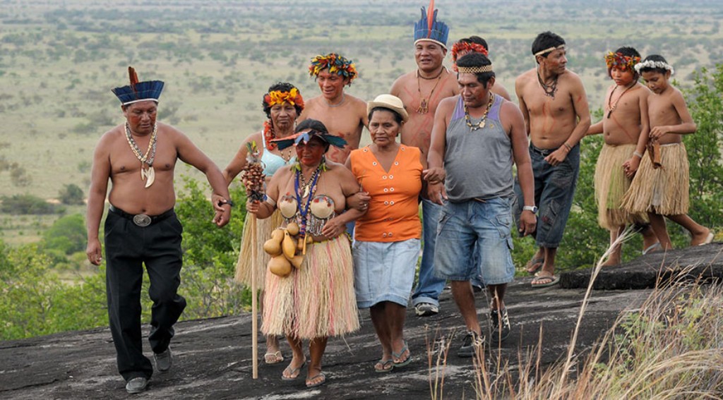 Grupo ind&iacute;gena em deslocamento sobre forma&ccedil;&atilde;o rochosa, com adornos, vestimentas e objetos tradicionais, em imagem que destaca a perman&ecirc;ncia de pr&aacute;ticas culturais, identidades coletivas e v&iacute;nculos com o territ&oacute;rio.IBGE.