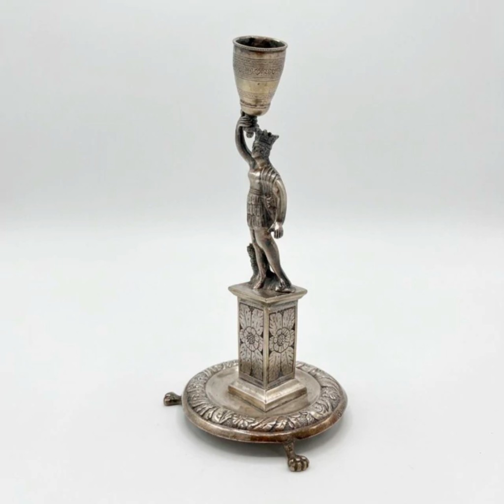 Raro paliteiro em prata de lei, executado pelas t&eacute;cnicas tradicionais da ourivesaria: batida e repuxada, em que a prata &eacute; modelada a martelo sobre moldes; e cinzelada, com ornamenta&ccedil;&atilde;o gravada &agrave; m&atilde;o; representando figura masculina coroada em armadura cl&aacute;ssica, erguendo com um bra&ccedil;o um recept&aacute;culo cil&iacute;ndrico para os palitos, sobre pedestal quadrangular com decora&ccedil;&atilde;o floral cinzelada.   A base circular moldurada com friso de folhas de acanto assenta sobre tr&ecirc;s p&eacute;s em garra de le&atilde;o, conferindo &agrave; pe&ccedil;a impon&ecirc;ncia e car&aacute;ter escult&oacute;rico. Apresenta contraste da cidade do Porto, Portugal, s&eacute;culo XIX.