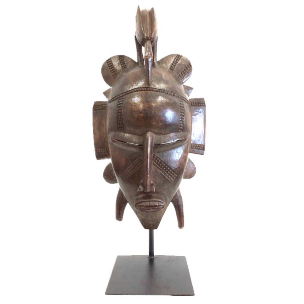 M&aacute;scara africana tribal esculpida em madeira, com rica ornamenta&ccedil;&atilde;o geom&eacute;trica e forte carga simb&oacute;lica ligada &agrave;s tradi&ccedil;&otilde;es espirituais e cerimoniais de povos da &Aacute;frica Ocidental. Utilizadas em rituais de inicia&ccedil;&atilde;o, celebra&ccedil;&otilde;es comunit&aacute;rias e cultos ancestrais, m&aacute;scaras como esta combinam express&atilde;o art&iacute;stica e fun&ccedil;&atilde;o ritual&iacute;stica, refletindo a profunda rela&ccedil;&atilde;o entre arte, religi&atilde;o e identidade cultural nas sociedades africanas tradicionais.