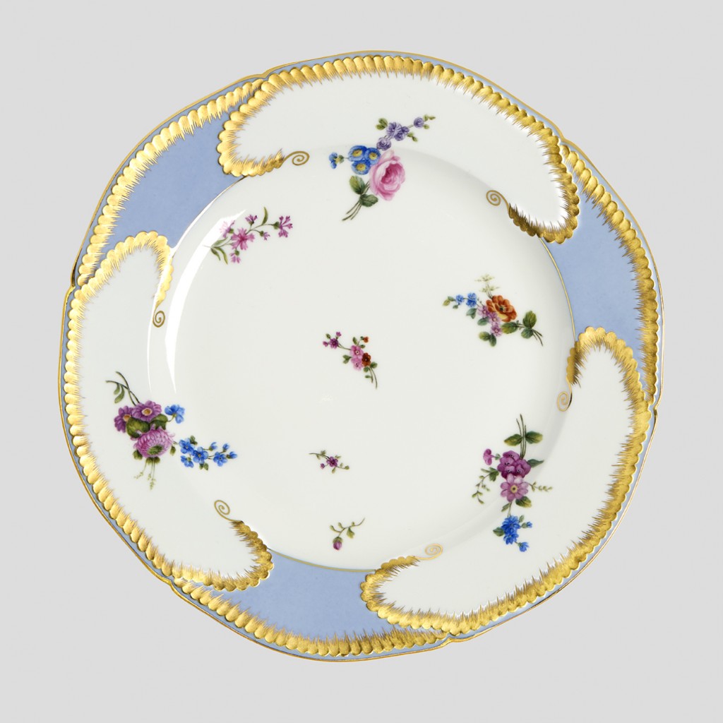 **Prato em porcelana de S&egrave;vres decorado com delicadas flores policromadas pintadas &agrave; m&atilde;o, borda em azul celeste e rica doura&ccedil;&atilde;o aplicada por artes&atilde;os especializados da manufatura francesa. Produzidas para atender aos padr&otilde;es de eleg&acirc;ncia da corte europeia do s&eacute;culo XVIII, pe&ccedil;as como esta exemplificam a combina&ccedil;&atilde;o entre sofistica&ccedil;&atilde;o t&eacute;cnica e sensibilidade art&iacute;stica que tornou S&egrave;vres uma refer&ecirc;ncia mundial nas artes decorativas. A leveza da composi&ccedil;&atilde;o floral, inspirada nos jardins e na bot&acirc;nica t&atilde;o apreciados pela aristocracia francesa, contrasta com a precis&atilde;o dos detalhes dourados e com a refinada harmonia crom&aacute;tica caracter&iacute;stica da manufatura. Objetos desse tipo integravam servi&ccedil;os de mesa destinados &agrave; nobreza e hoje s&atilde;o valorizados n&atilde;o apenas por sua beleza, mas tamb&eacute;m por seu papel na hist&oacute;ria da porcelana europeia e do gosto decorativo franc&ecirc;s.**