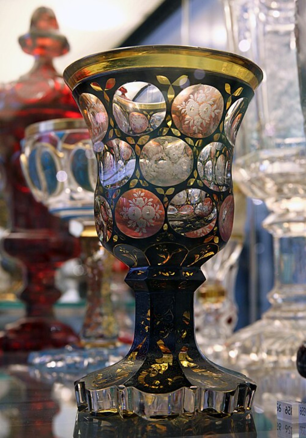 Exuberante exemplar da tradi&ccedil;&atilde;o vidreira da Bo&ecirc;mia, esta ta&ccedil;a combina cristal lapidado, detalhes dourados e reservas decorativas que revelam a extraordin&aacute;ria habilidade dos artes&atilde;os da Europa Central. Os medalh&otilde;es circulares ornamentados com motivos florais criam profundidade visual e valorizam os efeitos luminosos produzidos pelo cristal, enquanto a estrutura elegante da pe&ccedil;a evidencia a busca constante por refinamento est&eacute;tico. Obras desse tipo tornaram-se altamente apreciadas entre colecionadores e membros da aristocracia europeia, consolidando a reputa&ccedil;&atilde;o do Cristal Bo&ecirc;mia como uma das mais importantes express&otilde;es das artes decorativas ocidentais.