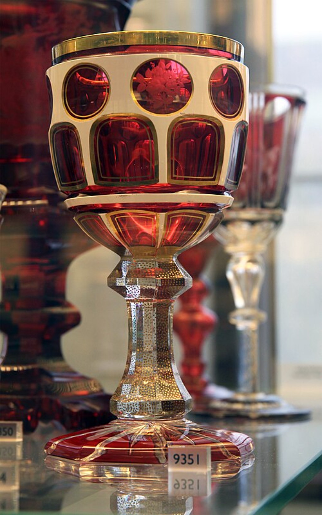 Exemplo not&aacute;vel da tradi&ccedil;&atilde;o vidreira da Bo&ecirc;mia, esta ta&ccedil;a em cristal rubi demonstra o elevado n&iacute;vel de especializa&ccedil;&atilde;o alcan&ccedil;ado pelas manufaturas da regi&atilde;o durante o s&eacute;culo XIX. A rica colora&ccedil;&atilde;o vermelha, obtida por meio da adi&ccedil;&atilde;o de elementos minerais &agrave; composi&ccedil;&atilde;o do vidro, contrasta harmoniosamente com as &aacute;reas lapidadas e transparentes, criando um refinado jogo de luz e profundidade. A ornamenta&ccedil;&atilde;o geom&eacute;trica evidencia a habilidade t&eacute;cnica necess&aacute;ria para trabalhar o cristal multicamadas, processo que exigia controle absoluto sobre o corte e o polimento da superf&iacute;cie. Obras como esta foram amplamente valorizadas por colecionadores e membros da aristocracia europeia, permanecendo at&eacute; hoje entre os exemplos mais admirados da arte decorativa produzida na Bo&ecirc;mia.