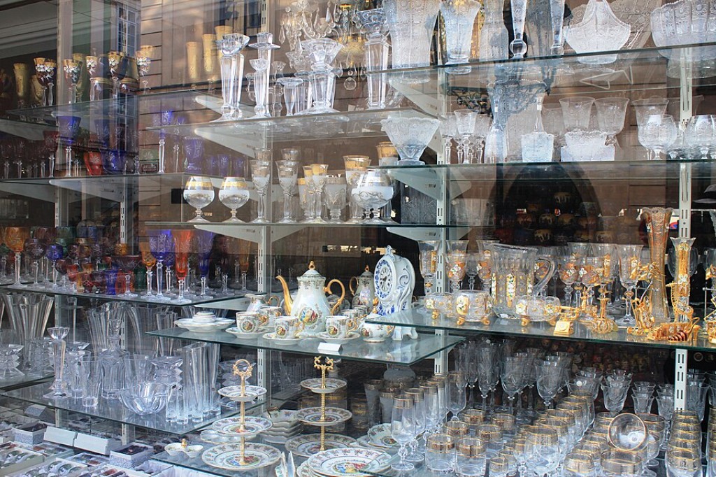 A ampla variedade de cristais expostos nesta vitrine ilustra a riqueza t&eacute;cnica e art&iacute;stica que caracteriza a produ&ccedil;&atilde;o vidreira europeia, especialmente a tradi&ccedil;&atilde;o bo&ecirc;mia. Ta&ccedil;as, vasos, licoreiras e objetos ornamentais convivem lado a lado, revelando diferentes estilos, per&iacute;odos hist&oacute;ricos e t&eacute;cnicas decorativas desenvolvidas ao longo dos s&eacute;culos. A presen&ccedil;a de pe&ccedil;as lapidadas, cristais coloridos e exemplares com detalhes dourados evidencia o elevado n&iacute;vel de sofistica&ccedil;&atilde;o alcan&ccedil;ado pelos artes&atilde;os da Bo&ecirc;mia, cuja influ&ecirc;ncia permanece viva no mercado de antiguidades e colecionismo. Ainda hoje, obras como essas s&atilde;o procuradas por sua beleza, raridade e import&acirc;ncia na hist&oacute;ria das artes decorativas ocidentais.