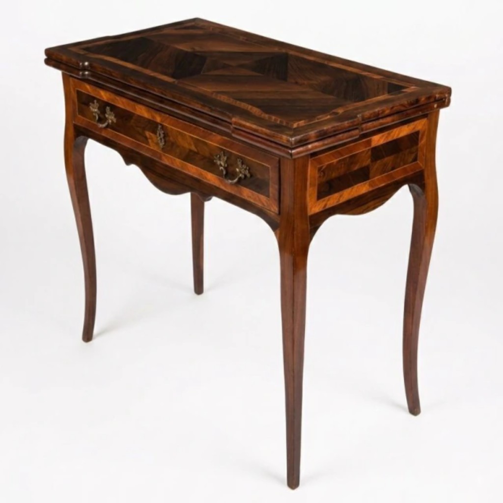 Mesa de jogo articulável executada em jacarandá, no estilo e época D. Maria I (1777-1816), com tampo retangular dobrável trabalhado em marchetaria de raios dispostos em composição geométrica simétrica — ao abrir, revela superfície interna revestida em feltro verde para uso em jogos de carta. Saia frontal com uma gaveta apresentando puxadores e espelhada em bronze cinzelado, e friso recortado em ondas. Apoia-se sobre quatro pernas em curva de delicada execução. Obra portuguesa do século XVIII/XIX, do período D. Maria I (1777-1816), de excelente qualidade.