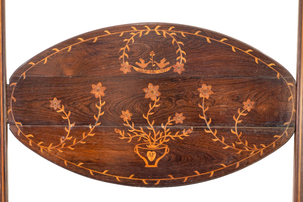 Detalhe da cabeceira de um leito de casal em jacarandá com marchetaria, produzido em Minas Gerais entre 1800 e 1825. Os delicados motivos florais e o lema "Amor nos Unio" evidenciam o refinamento da marcenaria brasileira do período colonial, que adaptou técnicas europeias às madeiras nobres nacionais.