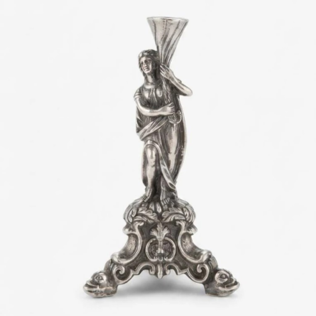 Paliteiro em prata de lei fundida e cinzelada, de produção brasileira do século XIX, com figura feminina alegórica em pé sustentando cornucópia perfurada para acomodação dos palitos, sobre base tripartida em volutas rocaille com apliques zoomorfos e florais. A peça integra a tradição da prataria brasileira oitocentista, na qual os paliteiros figurativos ganharam ampla difusão como peça de destaque das mesas postas do período imperial.