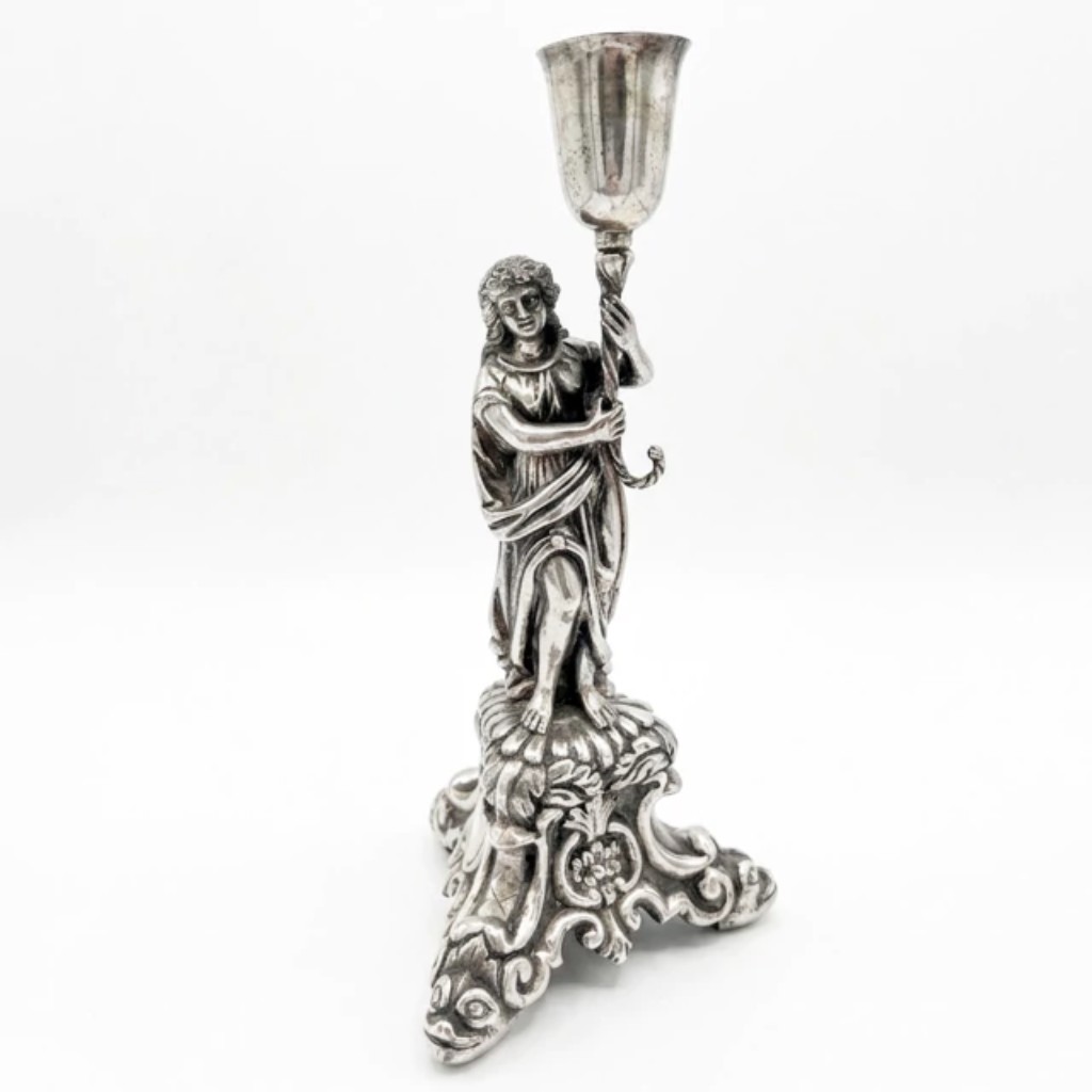 Raro paliteiro em prata de lei, executado pelas técnicas tradicionais da ourivesaria: batida e repuxada, em que a prata é modelada a martelo sobre moldes; e cinzelada, com ornamentação gravada à mão. Representando figura feminina em vestes clássicas com as mãos postas. A figura assenta sobre base com pés de cabeças de animais e ornamentação floral cinzelada, conferindo à peça marcado caráter de singular expressividade escultórica. Apresenta contraste da cidade do Porto. Portugal, século XIX.