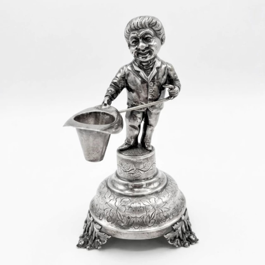 Raro paliteiro em prata de lei, representando figura masculina de expressão caricata, trajando casaco e segurando uma bengala e um chapéu cônico que serve de receptáculo para os palitos. Executado pelas técnicas tradicionais da ourivesaria: batida e repuxada, em que a prata é moldada a martelo sobre moldes; cinzelada, com decoração gravada à mão; e fundida nas partes escultóricas. A peça assenta sobre base abaulada com rica ornamentação floral e quatro pés em forma de folhagens, revelando o apuro artesanal da ourivesaria portuguesa oitocentista. Apresenta contraste. Portugal, século XIX.