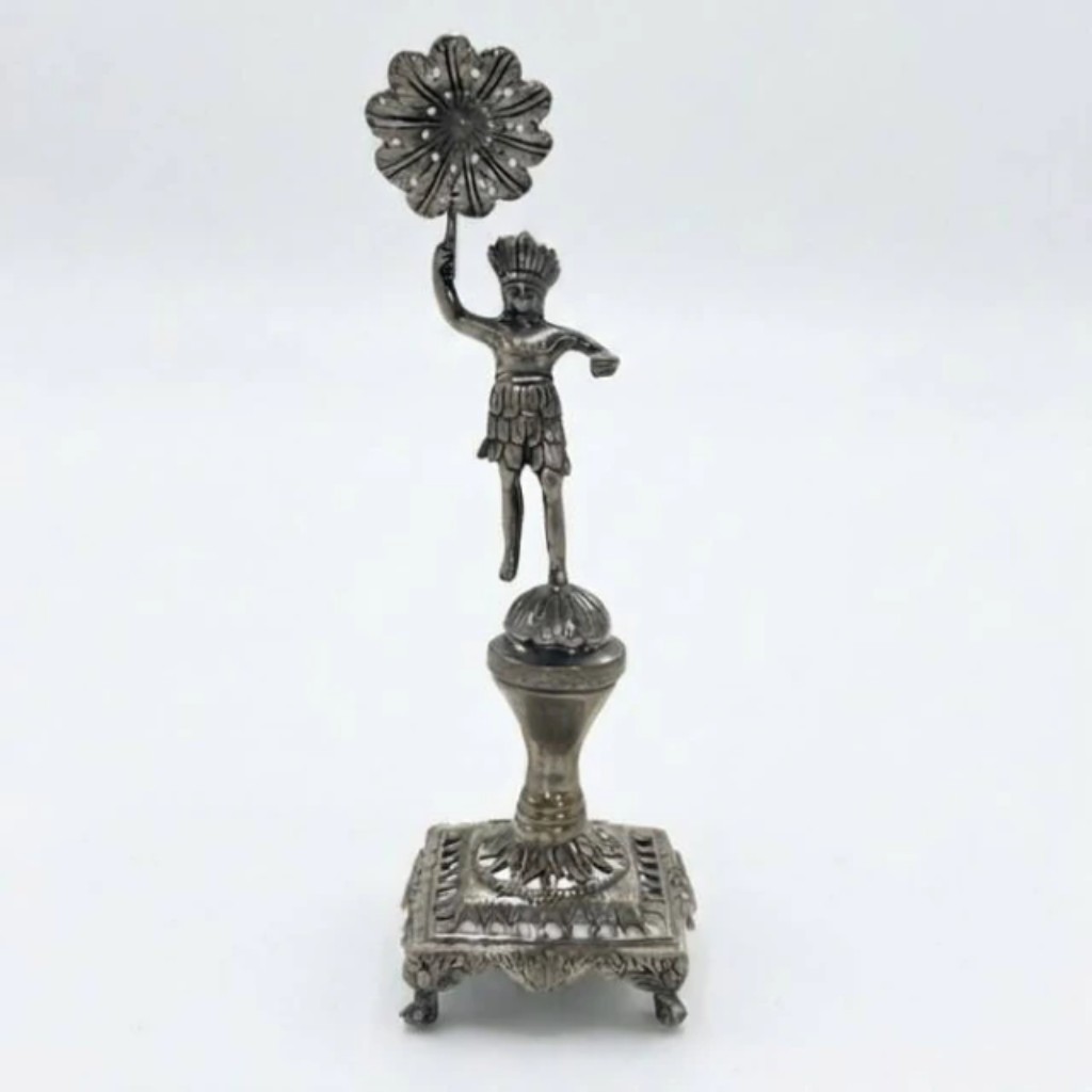 Paliteiro em prata de lei fundida e ricamente cinzelada, de produção brasileira do século XIX, com figura indígena central portando cocar e saiote de plumas sustentando roseta perfurada para acomodação dos palitos, sobre esfera de apoio.