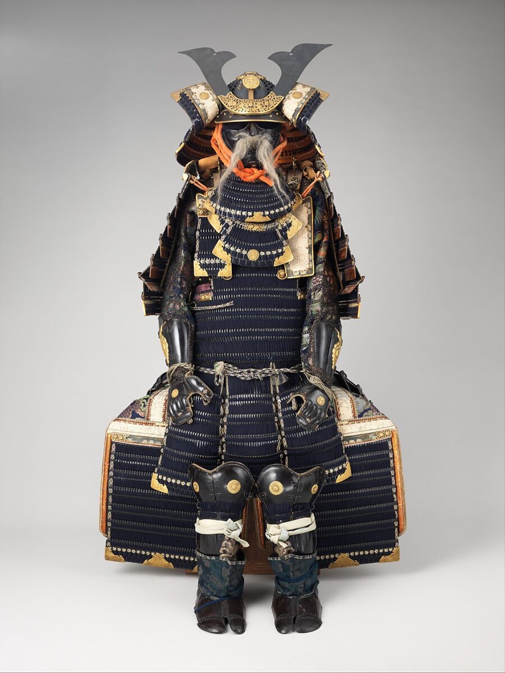 armadura japonesa do século XIX destaca-se pela sofisticação do seu trabalho em metal e detalhes ornamentais. O capacete e a máscara contêm inscrições do armeiro Jō Michitaka, revelando a autoria artesanal de alta elite da época. O conjunto traz o brasão (mon) de glicínia pertencente ao clã Ōkubo, que governava como daimyo (senhores feudais) no domínio de Karasuyama.