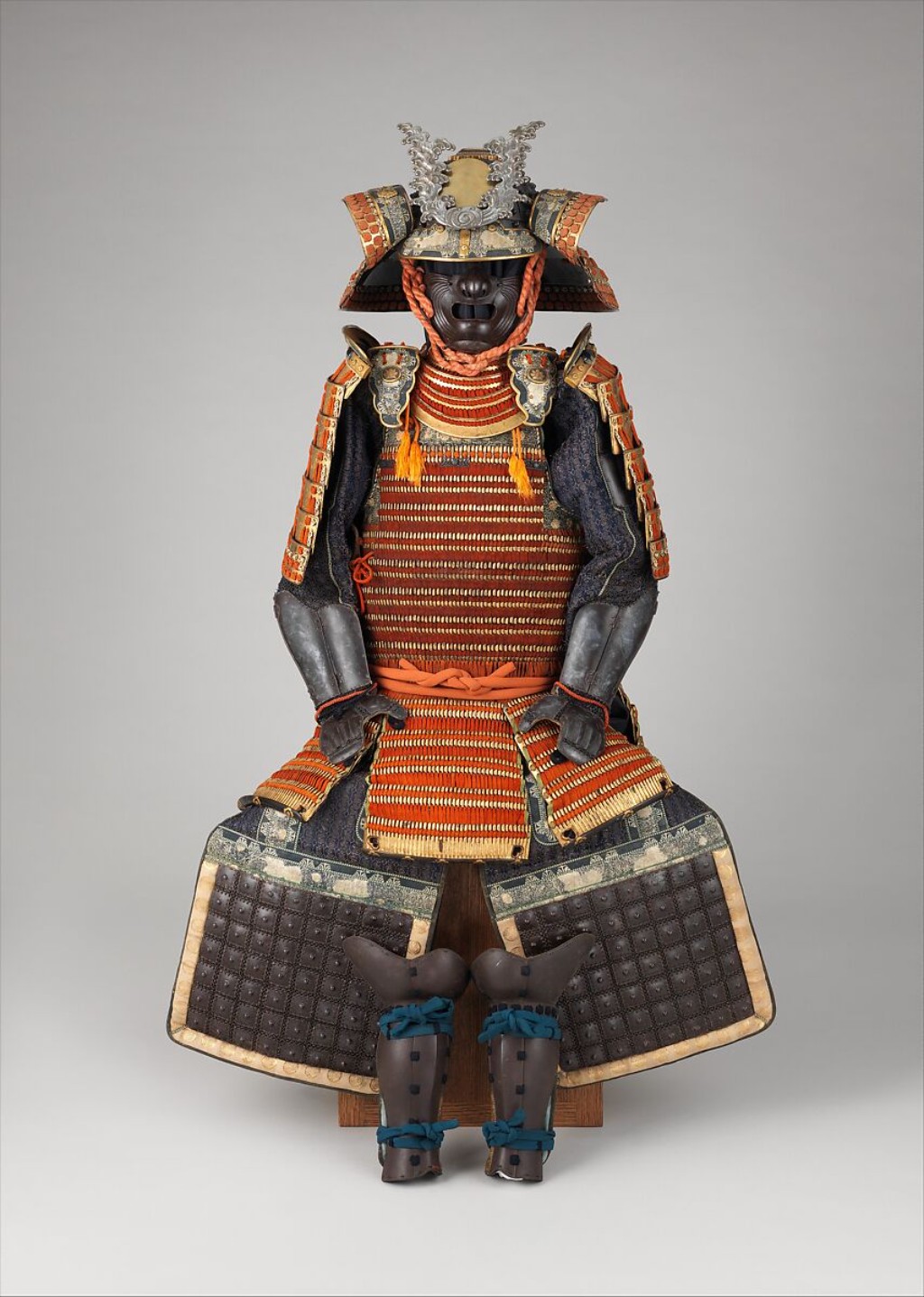 Armadura completa de samurai (yoroi) em tom alaranjado vibrante, apresentando intrincado trabalho de trançado de seda (odoshi) e placas de metal laqueado. O conjunto conta com capacete (kabuto) adornado com crista prateada (maedate), máscara facial feroz (menpo), protetores de ombro (sode), peitoral (dō) e caneleiras (suneate), exemplificando a suntuosidade da armaria japonesa do período feudal.