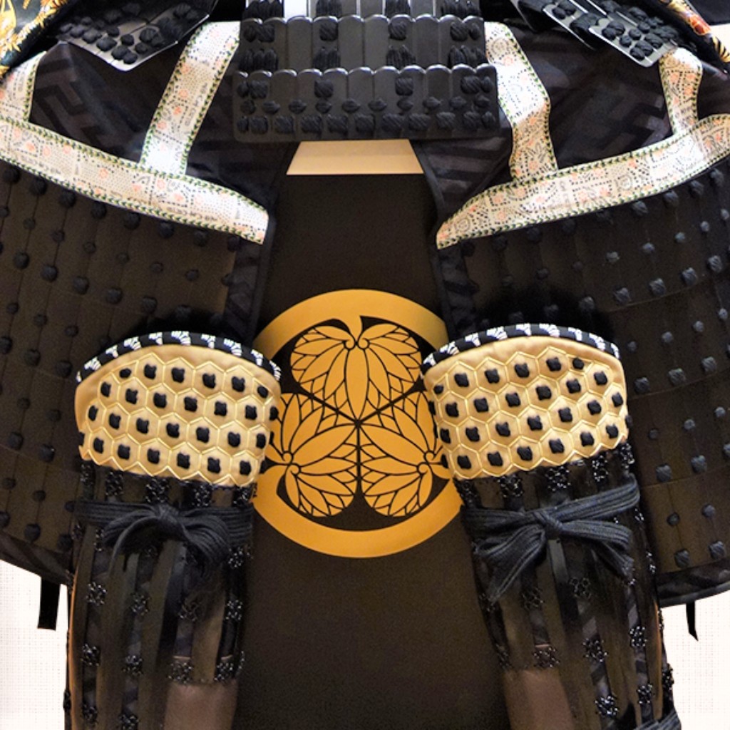 Detalhe de uma armadura de samurai do período Edo (1603–1868), exibindo o brasão Mitsuba Aoi do clã Tokugawa na couraça. Preservada no Museu de Arte Tokugawa, em Nagoya, a peça evidencia o refinamento da laca, da seda e da metalurgia que caracterizaram a armaria japonesa durante a era dos xogun