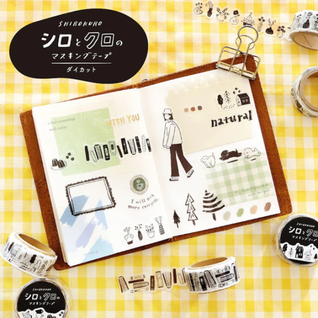 Vista superior de una libreta de viaje abierta sobre un mant&oacute;n a cuadros amarillos, decorada con cintas washi tape troqueladas e ilustraciones de la colecci&oacute;n Shirokuro en blanco y negro, acompa&ntilde;adas de stickers y rollos de washi tape a su alrededor.