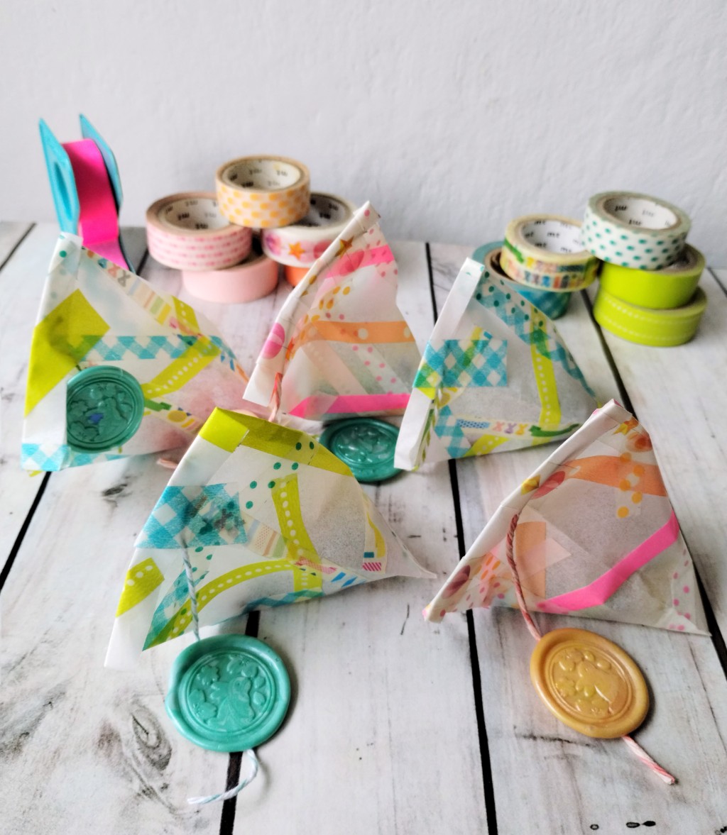 Washi tapes aplicadas a papeles de regalos:  Conjunto de sobrecitos de regalo con forma piramidal hechos de papel y decorados con tiritas de washi tape de colores vivos, acompa&ntilde;ados por varias cintas washi tape apiladas al fondo.