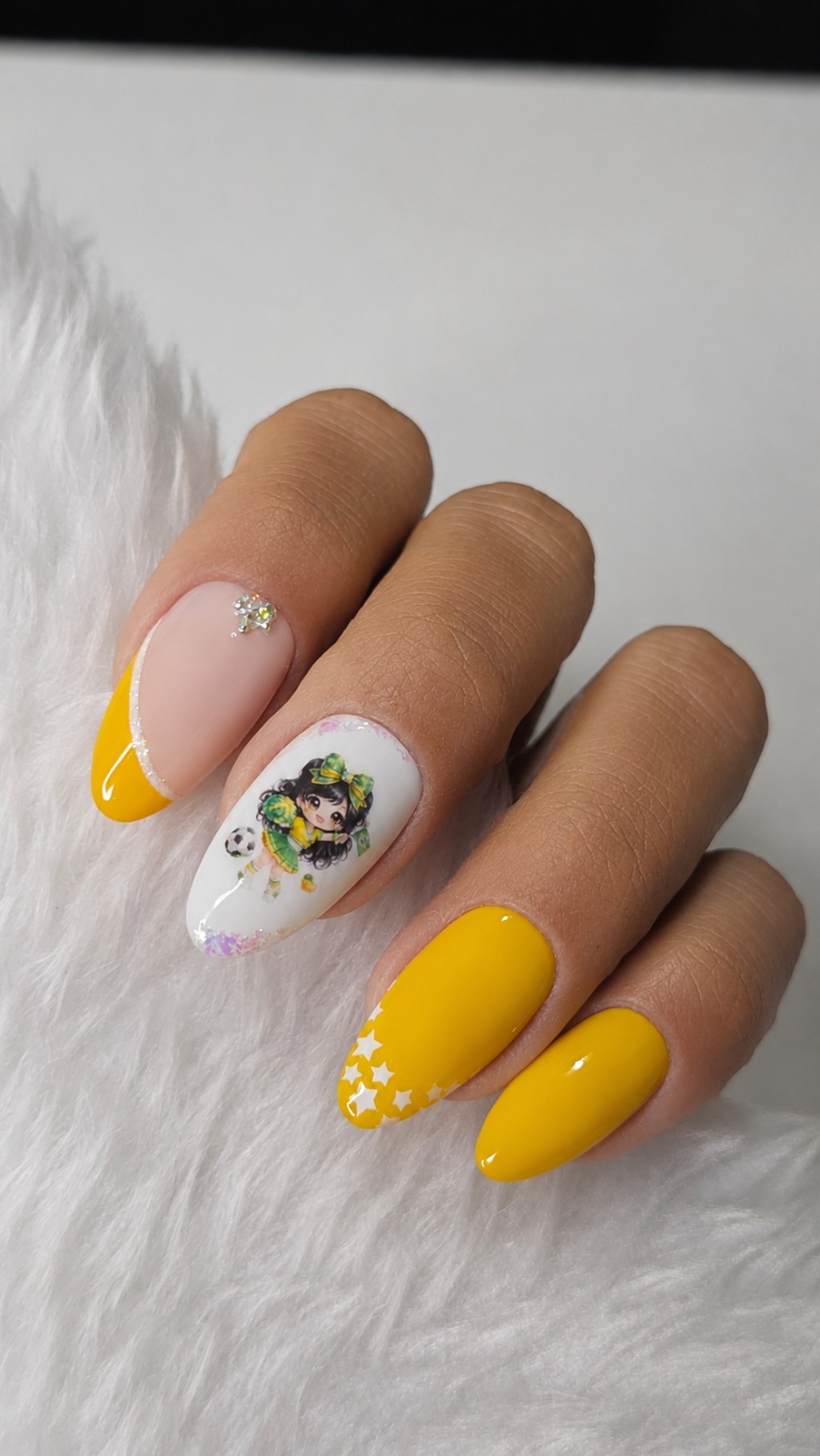 Unhas posti&ccedil;as decoradas em close com esmalta&ccedil;&atilde;o amarela brilhante, detalhe nude com strass e nail art feminina inspirada no Brasil.